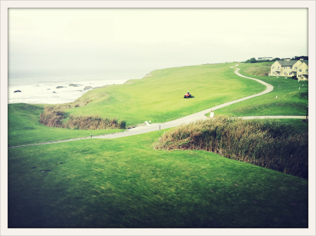 Eine Landschaftsszene mit Unkraut und Feldern im Vordergrund, einem Traktor und einem Weg in der Mitte, H├Ąusern und B├Ąumen oben rechts, Wasser oben links und einer bew├Âlktem Himmel.