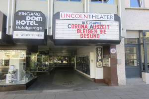 Das Lincoln Theater in Berlin, Deutschland ist ein Gebäude mit Glasfenstern und -türen, das eine Tafel mit Text und verschiedene Gegenstände im Inneren aufweist, die einen belebten Stadtbild-Eindruck vermitteln.