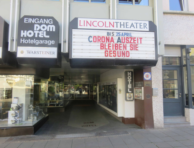 Das Lincoln Theater in Berlin, Deutschland ist ein Gebäude mit Glasfenstern und -türen, das eine Tafel mit Text und verschiedene Gegenstände im Inneren aufweist, die einen belebten Stadtbild-Eindruck vermitteln.