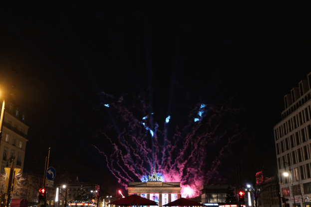 Eine belebte Stadtstraße in Berlin, Deutschland, mit dem Brandenburger Tor im Hintergrund, beleuchtet von Gebäudelichtern, Schildern, Verkehrsampeln, Zelten, Fahrzeugen und Feuerwerken am Nachthimmel.