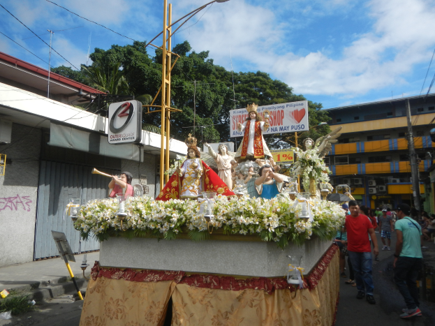 Ein Paradewagen mit Statuen, Blumen und Texttafeln, der eine Straße mit Laternenmasten, Kabeln, Gebäuden, Bäumen und einem bewölkten Himmel entlangfährt.