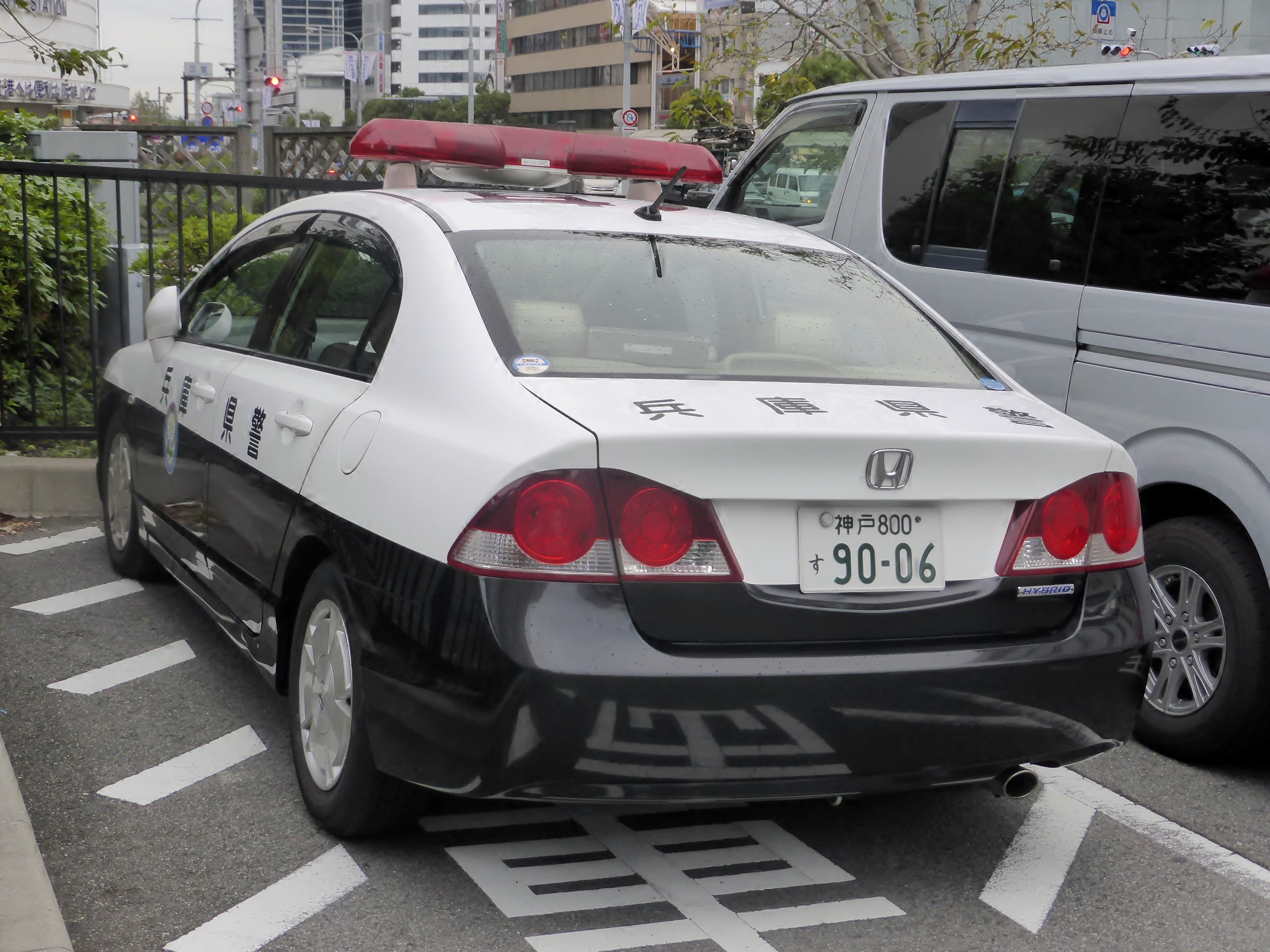 Polizeiauto neben einem weißen Lieferwagen auf einem eingezäunten Parkplatz mit Bäumen, Schildern, Straßenlaternen, Ampeln und Gebäuden unter einem bewölkten Himmel.