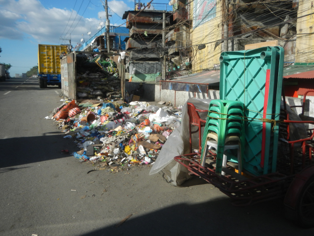 Ein Lastwagen neben einem Haufen Müll auf der Straße, mit einem Einkaufswagen mit Plastikstühlen rechts daneben und Gebäuden, Strommasten mit Drähten, Bäumen und einem bewölkten Himmel im Hintergrund.