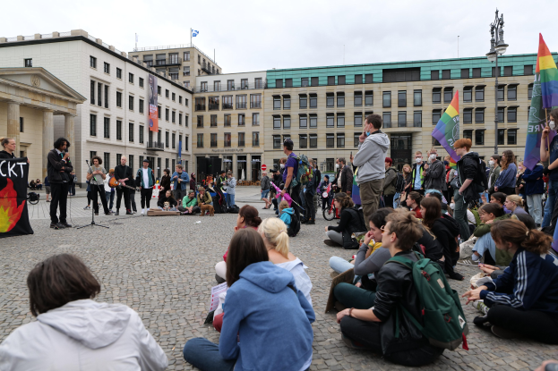 Gruppe von Menschen auf dem Boden sitzend vor einer gr"o\sseren Menge mit Fahnen, Spruchb"andern und einem Mikrofon auf einem St"and, mit einer Statue, Geb"auden und einem bew"olkten Himmel im Hintergrund bei einer Homo-Gegner-Demo in Berlin, Deutschland.