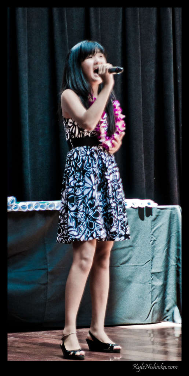 Eine Frau in einem bunten Kleid singt auf der Bühne in ein Mikrofon, mit einem Tisch voller Papiere und einem Vorhang im Hintergrund.