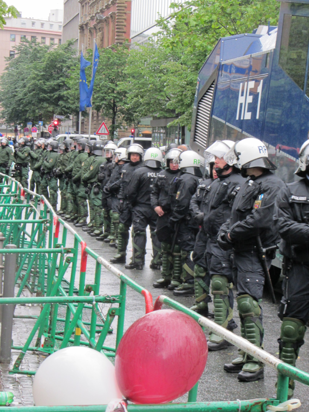 Gruppe von Polizeibeamten in Helmen und taktischer Ausrütständig hinter einer Barriere mit Geländern, Ballons auf der Straße und Gebäude, Flaggen und Bäume im Hintergrund unter einem klaren blauen Himmel.