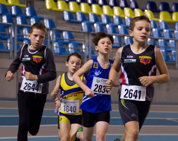 Vier Personen laufen, jede trägt ein Kleid in einer anderen Farbe mit einem Aufkleber, blaue und gelbe Stühle sind im Hintergrund zu sehen.