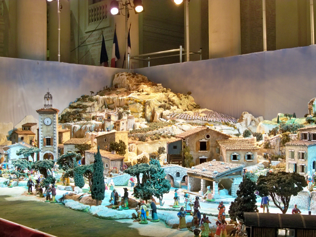 Eine Museumsausstellung eines Krippenmodells mit Menschen, Häusern, Bäumen, Steinen, einem Glockenturm, Fahnen, Pfählen und Lichtern.