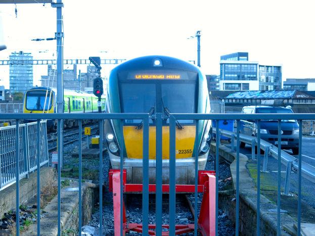 Zwei Züge auf dem Gleis mit einem Zaun im Vordergrund, Pfosten auf dem Land, einer mit einer Signalleuchte, ein Auto auf der Straße rechts, Gebäude im Hintergrund und den Himmel oben.