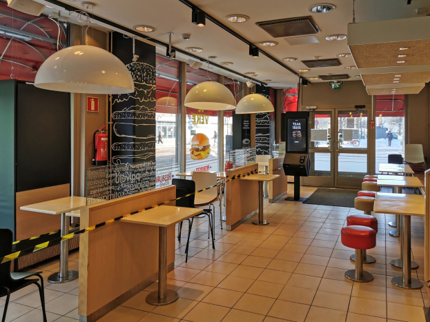 Innenraum eines Schnellrestaurants mit Tischen, Stühlen, Hockern, einem Boden, Deckenleuchten, einem wandmontierten Feuerlöscher, Glastüren und weiterer Einrichtung.