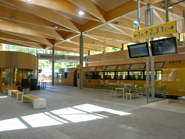 Ein belebter Bahnhof mit einem Zug auf den Schienen, umgeben von Bänken, Tischen, Pfosten, Schildern, Laternen und Bäumen, wo Menschen auf den Zug warten.