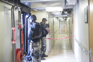 Zwei Polizisten in Uniform stehen nebeneinander in einem Flur, tragen Helme und halten Waffen, mit einer Tür und einem Foto auf der rechten Wand und Deckenleuchten und Rohren oben.