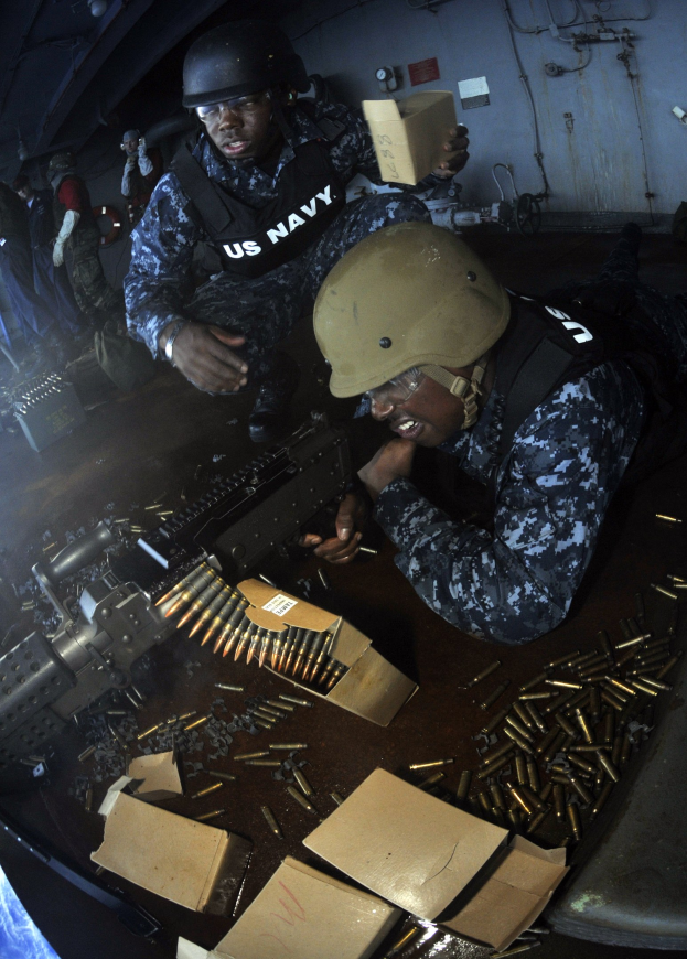 Zwei Militärangehörige in Uniform und Helmen, die eine Maschinenpistole handhaben, umgeben von verstreuter Munition und Ausrüstung, mit einer Wand aus Rohren und diversen Gegenständen im Hintergrund.