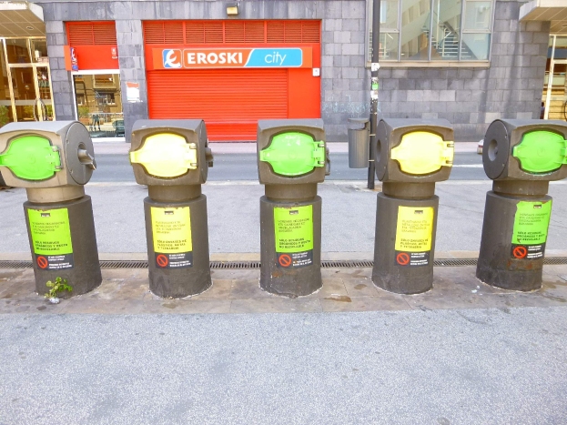 Eine Reihe von Parkautomaten entlang einer Straße mit einem Gebäude mit Glasfenstern und einer Rolllade mit Text sowie einem nahen Müllcontainer.