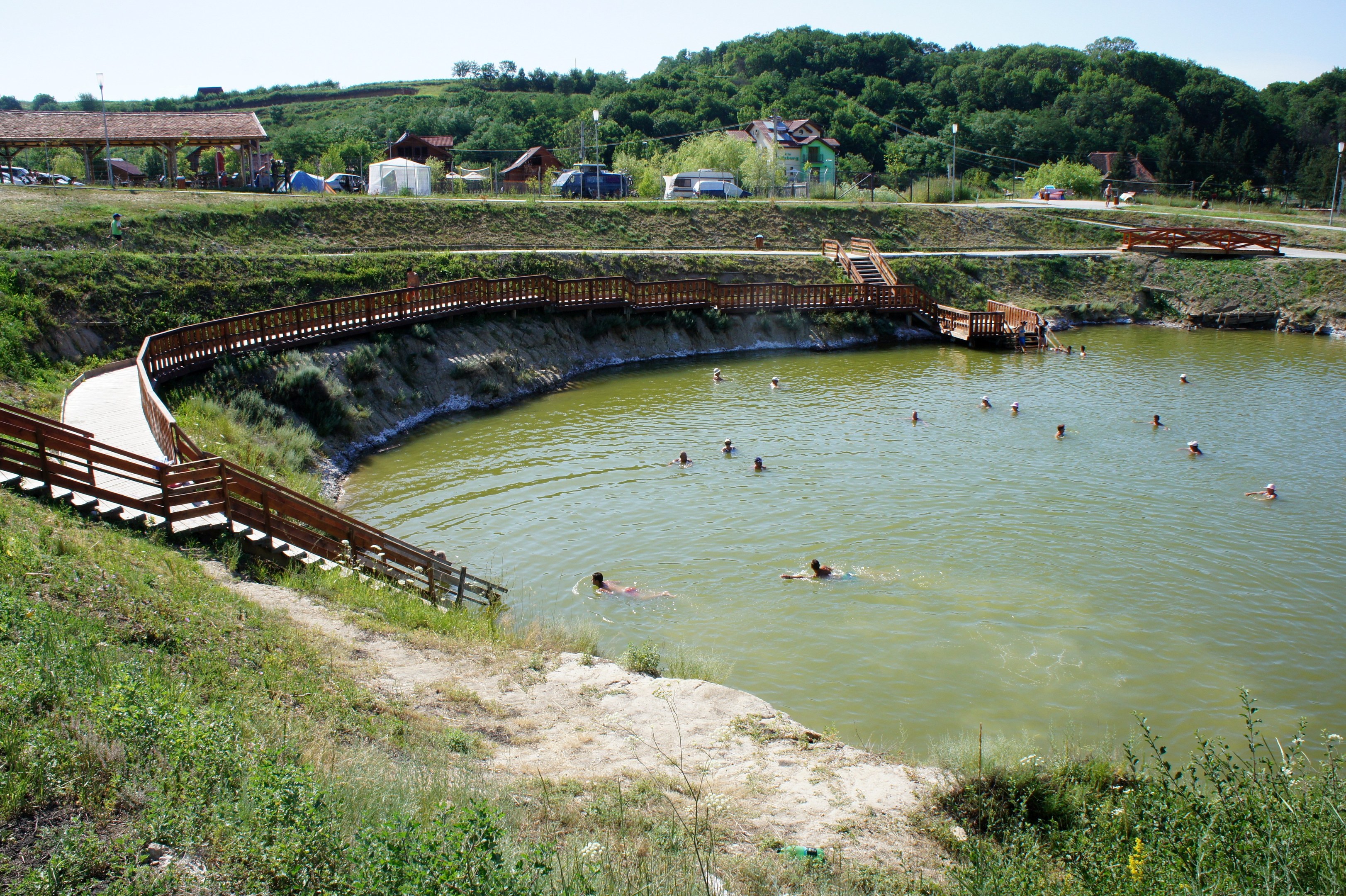 Menschen schwimmen in einem Gewässer umgeben von Grün, mit einer Brücke, Treppen, Hütten, Fahrzeugen, Pfählen und einem klaren blauen Himmel im Hintergrund.