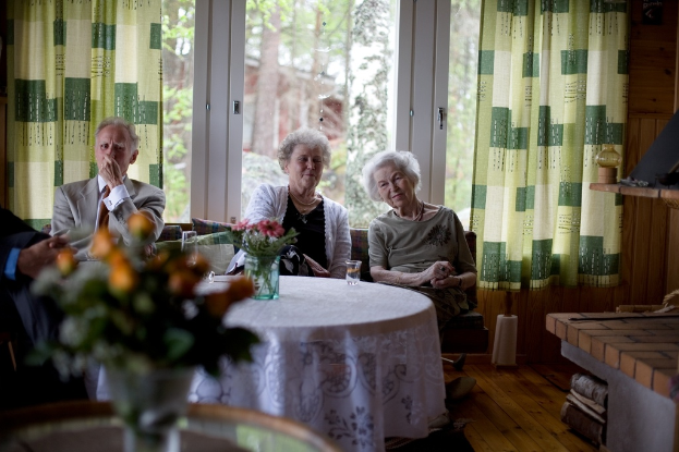 Eine Gruppe älterer Personen, die auf einem Sofa sitzen und vor sich einen Tisch mit einer Vase darauf haben. Im Hintergrund gibt es eine Glastür und Vorhänge.