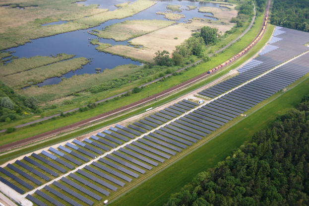 Luftaufnahme einer Solar-Farm mit Solar-Panelen, einem Zug auf einem Bahngleis, umgeben von Bäumen, Gras, Pflanzen und Wasser.