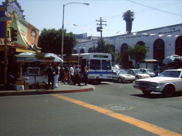 Eine belebte Straßenszene mit mehreren Fahrzeugen, Gebäuden auf beiden Seiten, einem Stand auf der linken Seite, Bäumen auf der rechten Seite und einem Himmel darüber.