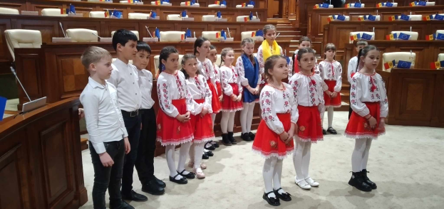 Eine Gruppe von Kindern in weißen und roten Kleidern steht in einem Raum mit einem Podium, Mikrofon, Flaggen, Stühlen und Tischen im Hintergrund.