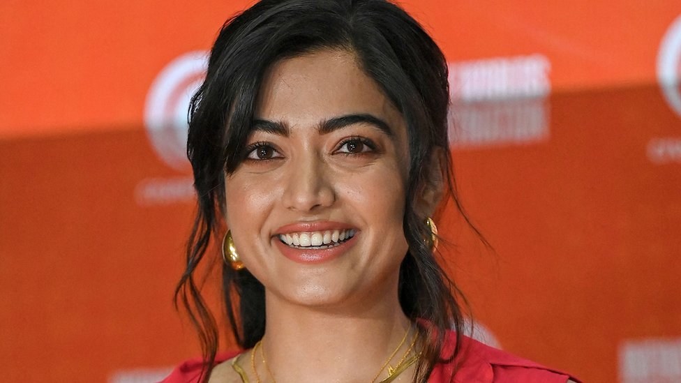Eine Frau in einem roten Kleid mit einem eleganten Dutt und dezentem Make-up steht strahlend da, trägt schwarze High Heels und hat ein hellgelbes Banner mit der Aufschrift "Sonam Kapoors beste Schönheits Looks vom Cannes Film Festival" im Hintergrund.