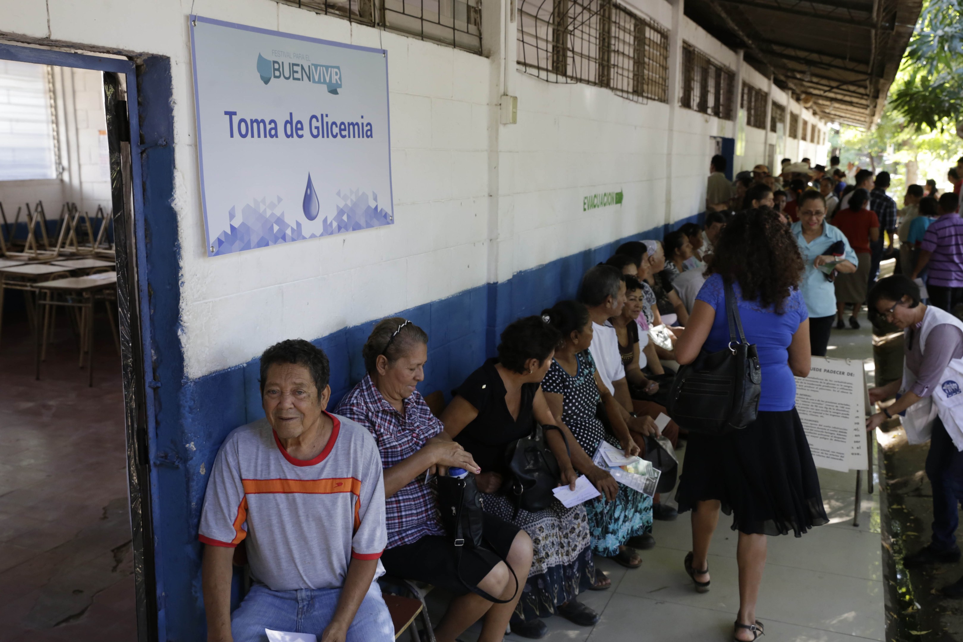 Menschen sitzen auf Bänken vor einem Gebäude, einige halten Papiere und Taschen, mit einer Tafel an der Wand, Tischen und Stühlen links daneben und Bäumen und einer Hütte im Hintergrund.