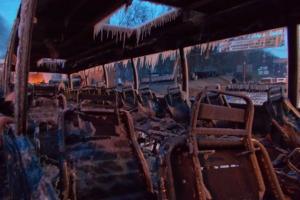 Ein zerstörter Bus mit verkohlten Sitzen und Eiszapfen, die von der Decke hängen, vor dem Hintergrund von Bäumen, Gebäuden und einem klaren Himmel.