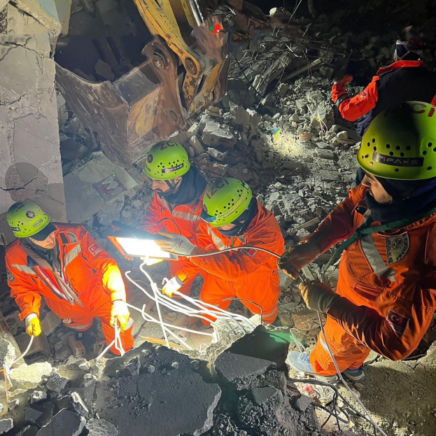 Eine Gruppe von Rettungskräften in orangefarbenen Uniformen und grünen Helmen beseitigt Trömer von einem eingestürzten Gebäude mit einem Kran im Hintergrund.