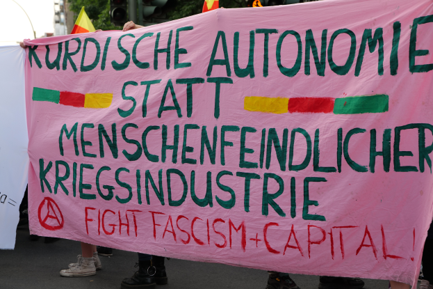 Eine Gruppe von Menschen auf einer Straße mit einer pinken Fahne, auf der "Kurdische Autonomie Statt Menschenfeindlicher Kriegsindustrie Fight Fascism Capital" steht, mit Ampeln, Flaggen, Bäumen, Gebäuden und einem klaren blauen Himmel im Hintergrund.