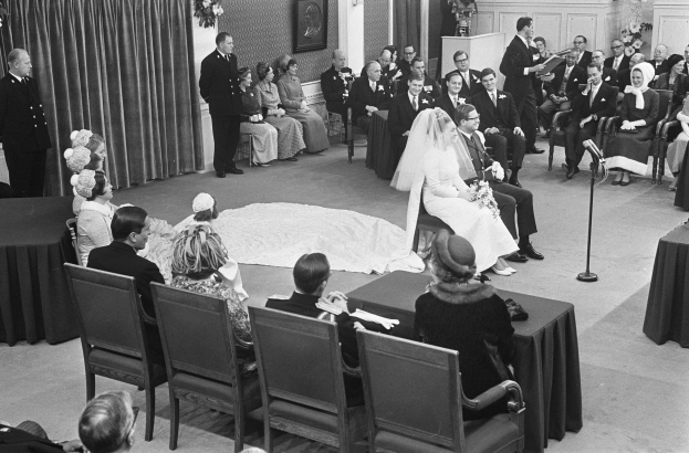 Schwarzes und weißes Foto einer Hochzeitszeremonie mit sitzenden Gästen, einem Mann mit Mikrofon, einer Frau mit Blumenstrauß, einem Podium mit Mikrofon, Blumenschmuck, einem Vorhang, einem Rahmen an der Wand und einer Tür im Hintergrund.