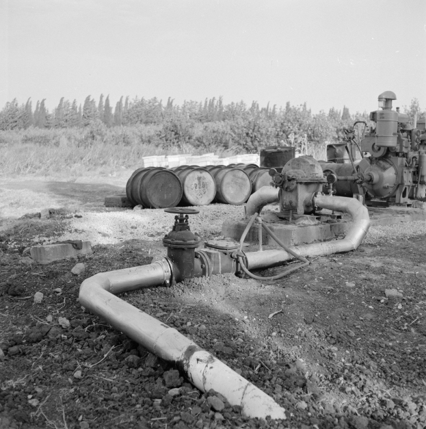 Ein Schwarz-Weiß-Foto einer Gasleitung in einem Feld, umgeben von Maschinen, Rohren und Fässern, mit Bäumen und einem klaren Himmel im Hintergrund.