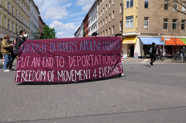 Eine Gruppe von Menschen marschiert auf einer Stadtstraße und trägt ein Banner, auf dem die Öffnung der Grenzen und das Ende der Abschiebungen gefordert wird, mit Gebäuden, Bäumen und Fahrrädern an der Straße und einem bewölkten Himmel.