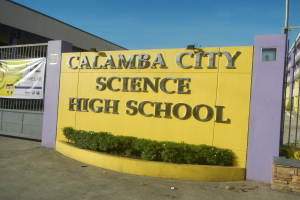 Gebäude der Calamba City Science High School mit einem Namensschild, Pflanzen, einer Fahne, einem Tor, einem Pfosten und einem klaren blauen Himmel im Hintergrund.
