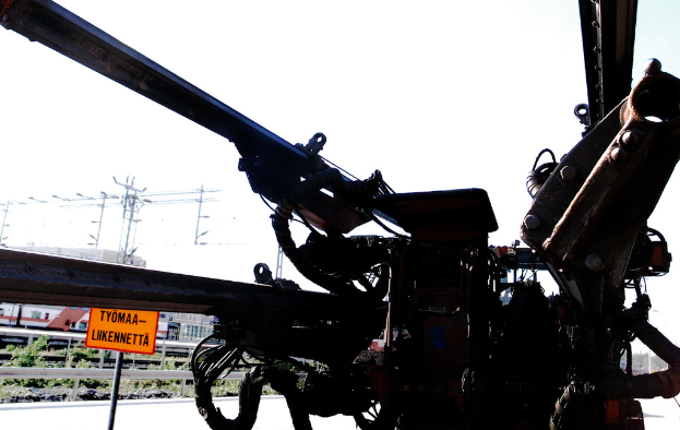 Eine Maschine im Vordergrund mit Strommasten, Kabeln, Barrieren, Pflanzen und Himmel im Hintergrund.