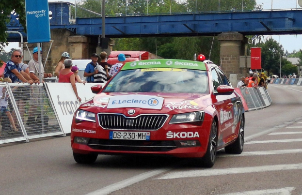 Roter Wagen fährt an einer Menschenmenge vorbei auf einer Straße mit Bäumen, Geländern, Bannern, Laternenmasten und einer Brücke im Hintergrund bei bewölktem Himmel.