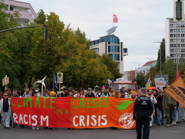 Eine Gruppe von Menschen marschiert eine von Bäumen gesäumte Straße entlang und hält ein Banner in die Höhe, auf dem 'Klimakrise ist eine Krise' steht, mit Gebäuden und einem klaren blauen Himmel im Hintergrund.