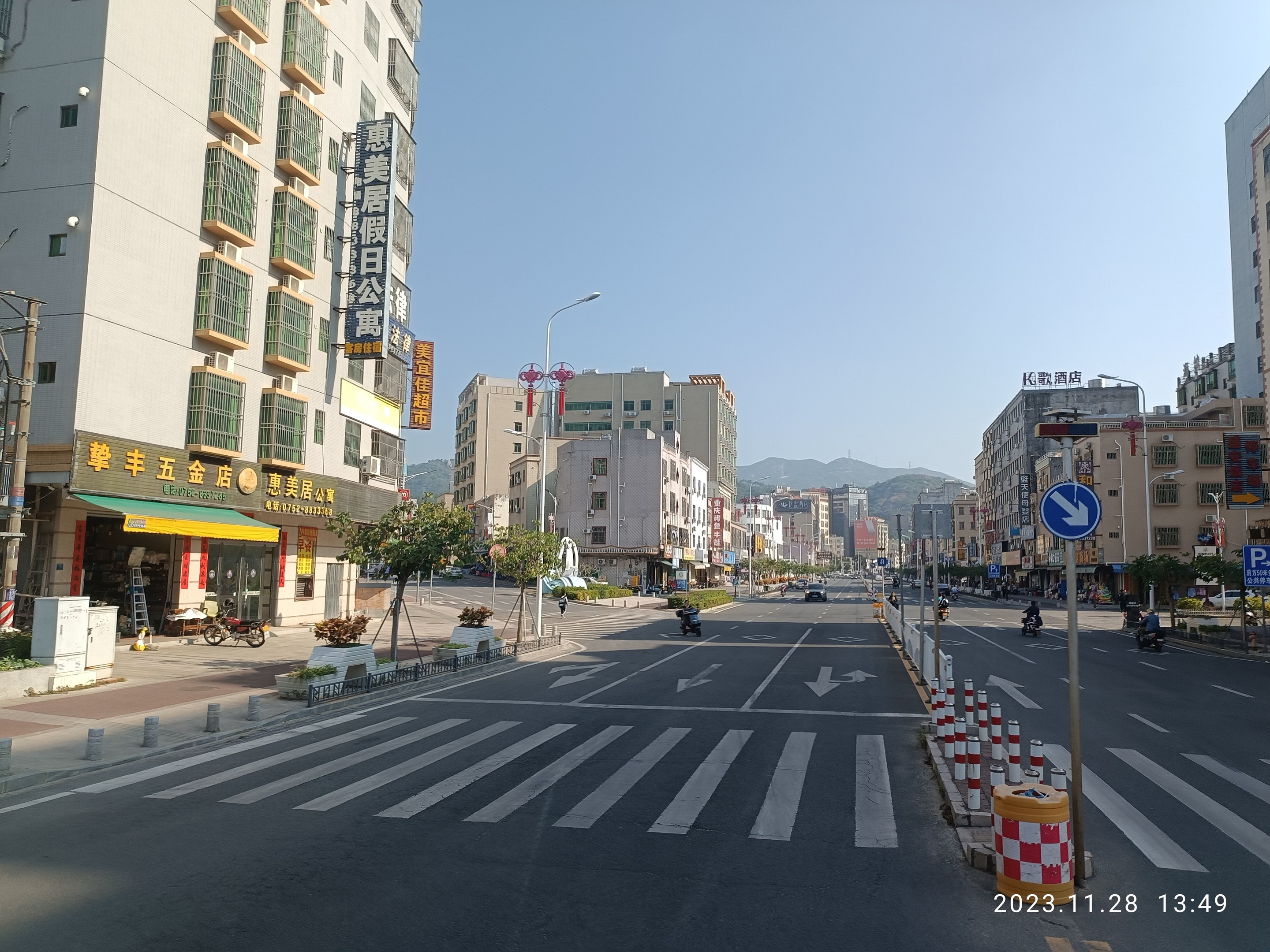 Eine Stadtstraße mit Gebäuden, Straßenlaternen, Verkehrszeichen, Kraftfahrzeugen, Barrieren, Bäumen, Pflanzen, Namensschildern, Strommasten, Stromkabeln, Hügeln und Himmel im Hintergrund.