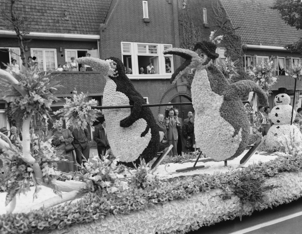 Schwarzes und weißes Foto eines Umzugswagens, der mit Blumen und Pinguinen geschmückt ist, mit Menschen auf der Straße und Gebäuden mit Fenstern im Hintergrund.