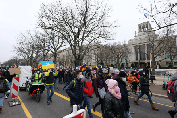 Eine große Gruppe von Menschen marschiert auf einer Straße in Washington, D.C., mit Schildern und Transparenten, einige fahren Fahrräder, unter einem klaren blauen Himmel vor einem Gebäude mit Schildern und Bäumen.