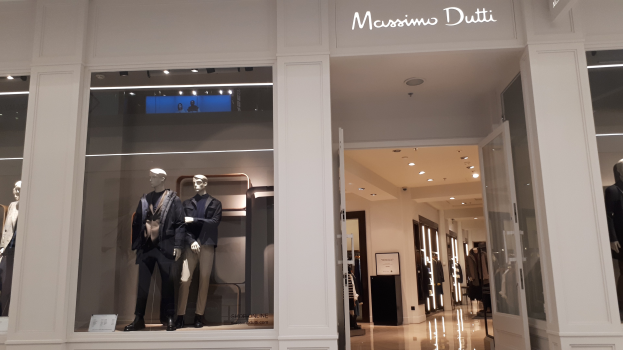Vitrine des Massimo Dutti in Mailand mit Schaufensterpuppen in der aktuellen Mode, beleuchteter Decke, Informationsschildern und sichtbarem Boden.