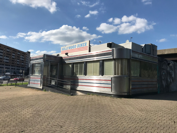 Außenansicht des Hollywood Diners mit seiner Leuchtschrift, das sich auf einer Straße befindet, die von Gebäuden, Straßenmöbeln und Fahrzeugen gesäumt ist.