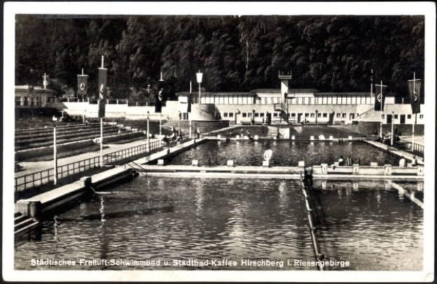 Schwarzes und weißes Foto eines Schwimmbads mit Gebäuden, Pfählen, Flaggen, Treppen, Geländern und Bäumen drumherum, mit Text unten.