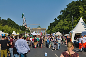 Eine Menschenmenge, die eine Straße entlanggeht, die mit Zelten, Fahrzeugen und Bäumen gesäumt ist, mit einem Bogen und einem klaren blauen Himmel im Hintergrund und Polen mit Fahnen auf der linken Seite, die wahrscheinlich das Oktoberfest in München, Deutschland, darstellen.