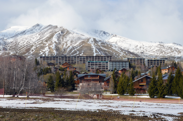 Ein Skigebiet mit schneebedeckten Bergen im Hintergrund, schneebedeckter Boden mit Gras- und Pflanzenflecken, Bäume und Gebäude im Vordergrund und eine bewölkte Himmelslandschaft.