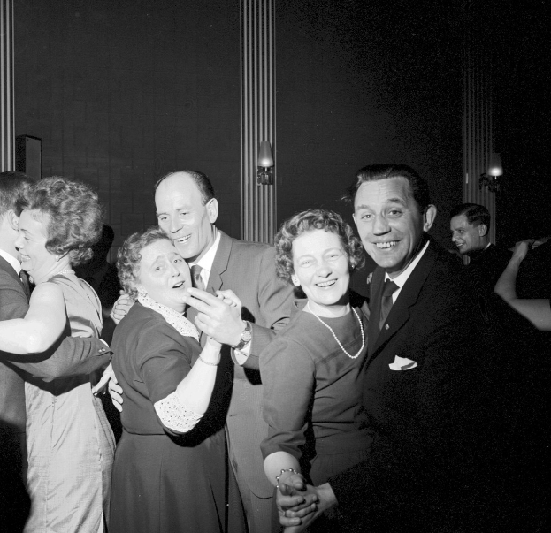 Schwarzes und weißes Foto einer Gruppe von Menschen, die zusammen auf einer Party tanzen, sich an den H├Ąnden halten und l├Ącheln, mit beleuchteter Wand im Hintergrund.