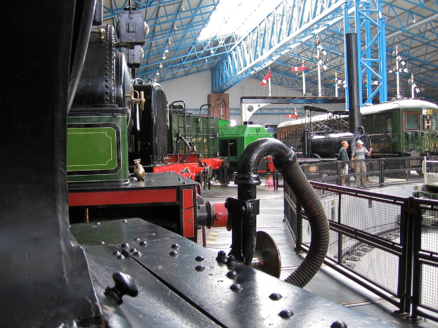 Mehrere Lokomotiven sind in einem Abstellgleis mit zahlreichen Eisenstangen darüber, einem Zaun auf der rechten Seite, einem Zug auf dem Bahnsteig neben dem Zaun und zwei Personen in der Mitte neben dem Zaun geparkt.