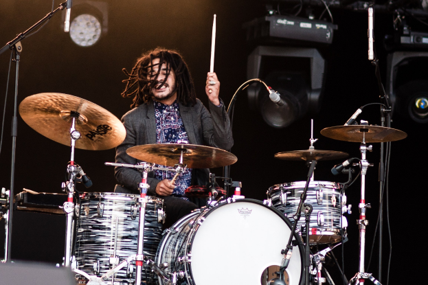 Ein Mann mit Dreadlocks, der auf der Bühne Schlagzeug spielt, hält Drumsticks in der Hand und hat Mikrofonständer vor sich, vor einem dunklen Hintergrund mit Bühnenbeleuchtung.