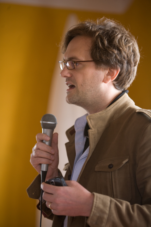 Ein Mann in einer Jacke hält ein Mikrofon, spricht und schaut nach links, mit einer Wand und einem weißen Tuch im Hintergrund.