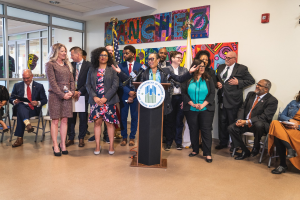 Gruppe von Menschen in einem Raum mit einem Podium, einem Mikrofon und einer Tafel mit der Aufschrift "Sancho-Stadtrat tritt dem Stadtrat bei", umgeben von Flaggen, Stühlen und einem Feuerlöscher.