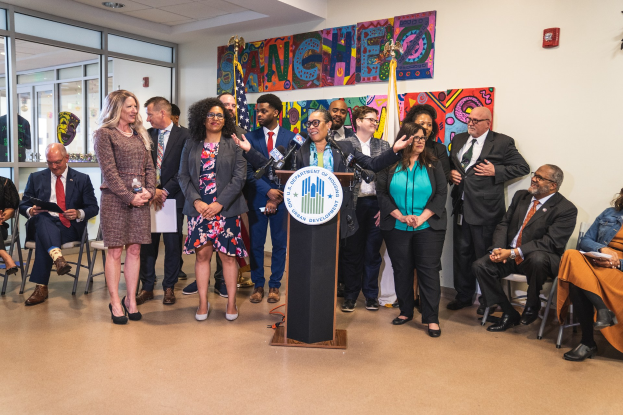 Gruppe von Menschen in einem Raum mit einem Podium, einem Mikrofon und einer Tafel mit der Aufschrift "Sancho-Stadtrat tritt dem Stadtrat bei", umgeben von Flaggen, Stühlen und einem Feuerlöscher.