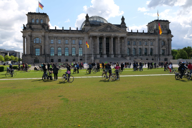 Menschen auf Fahrrädern vor dem Reichstaggebäude in Berlin, Deutschland, mit Bäumen, Pflanzen und einer bewölkten Himmel.
